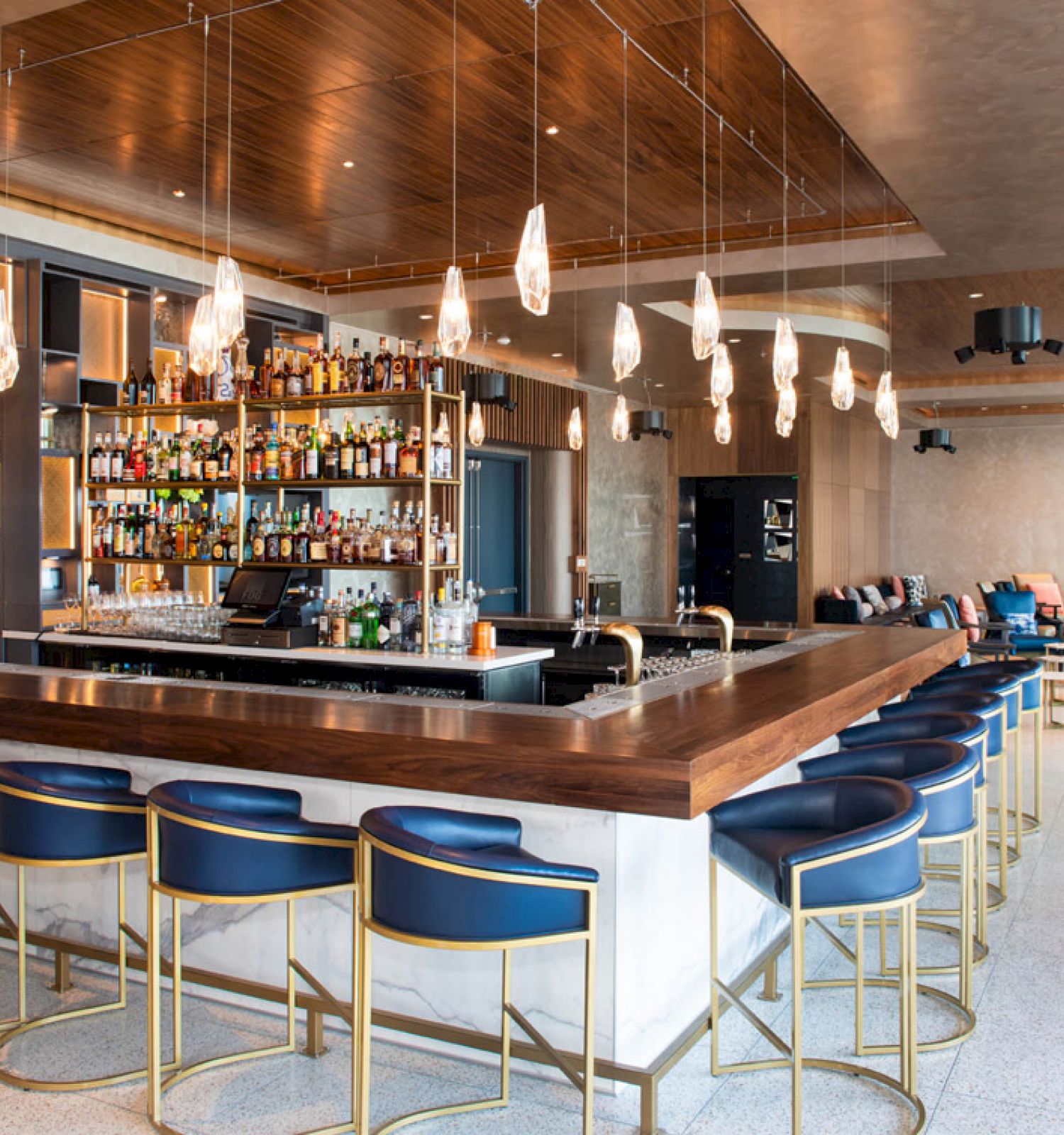A modern bar with blue seats, pendant lights, and a well-stocked liquor shelf. The area has a cozy seating section and a contemporary design.
