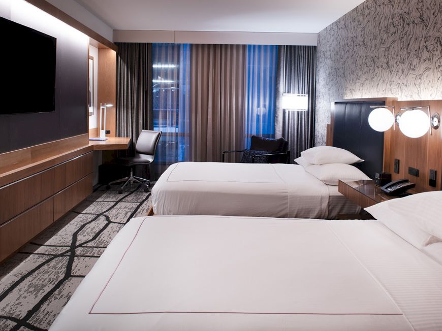 A modern hotel room with two double beds, a wall-mounted TV, a desk with a chair, stylish lighting, and large windows covered by curtains.
