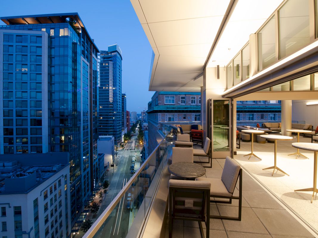 A modern cityscape at dusk with tall buildings. A partially enclosed rooftop terrace displays dining tables, lounge chairs, and city views.