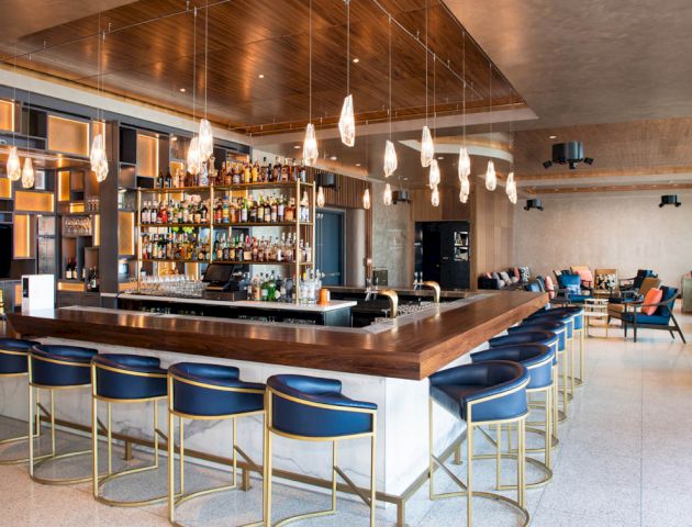 The image shows a modern bar with a wooden counter, blue and gold barstools, hanging light fixtures, and a well-stocked backbar.