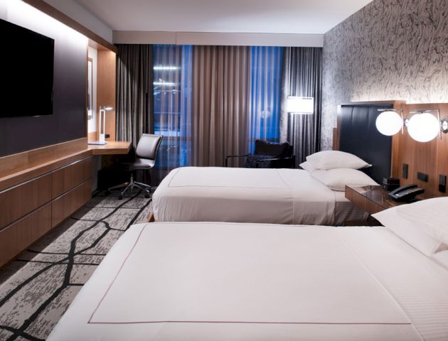 A modern hotel room featuring two neatly made double beds, a wall-mounted TV, a work desk with a chair, stylish decor, and adequate lighting.