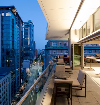 A modern rooftop patio with seating overlooks a cityscape of high-rise buildings at dusk, with the street below illuminated by city lights.