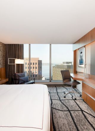 A modern hotel room features a large bed, a wall-mounted TV, a desk with a chair, and a cityscape view through the window.