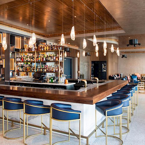 A modern bar with a wooden counter, blue barstools, hanging lights, and a fully stocked bar in the background. Lounge seating is visible further back.