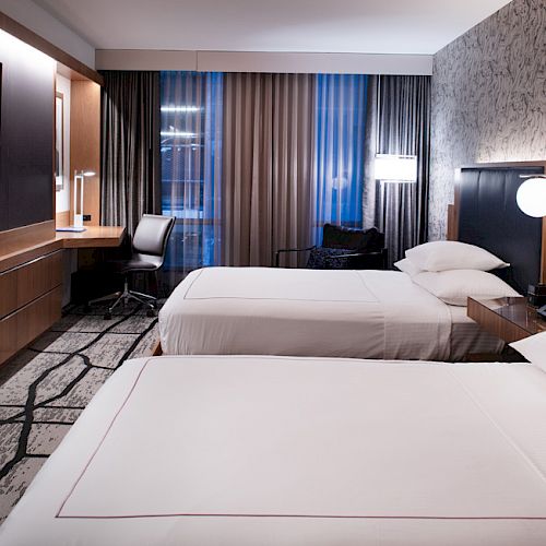 A modern hotel room with two beds, a wall-mounted TV, a desk, and lamps. The room features patterned wallpaper and floor-to-ceiling windows.