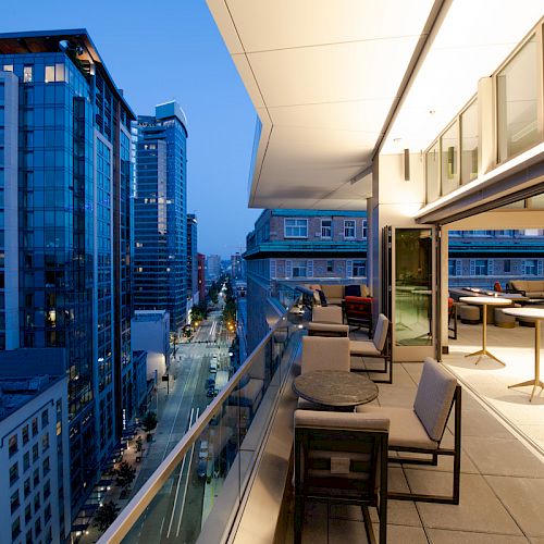 The image shows a modern cityscape with tall buildings at dusk, viewed from a high-rise balcony furnished with tables and chairs.