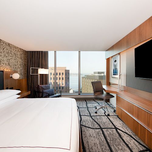 The image shows a modern hotel room with a large bed, desk, chair, wall-mounted TV, and a window offering a city view.