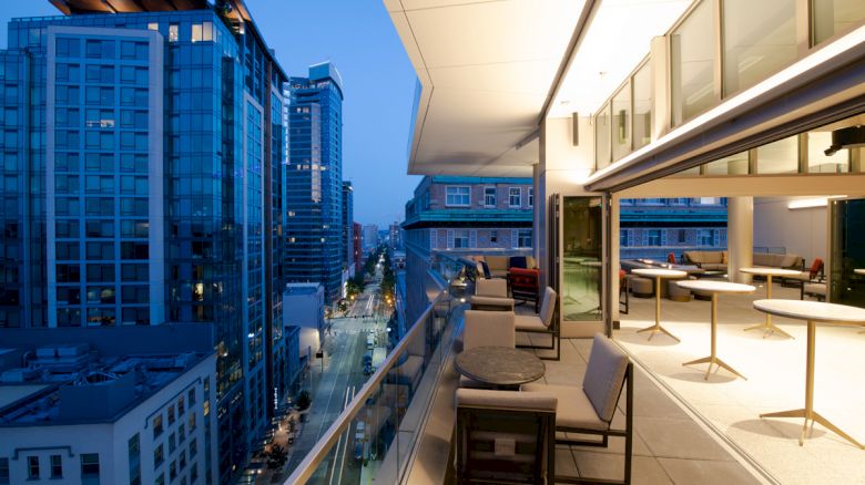 An elegant balcony with seating and tables overlooks a city street, surrounded by modern skyscrapers in the evening light.
