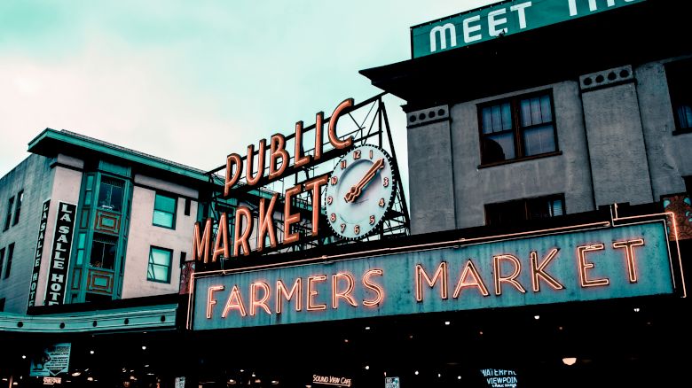 An outdoor market sign reads "PUBLIC MARKET" and below it, "FARMERS MARKET." Visible buildings include a hotel; part of another sign reads "MEET THE."