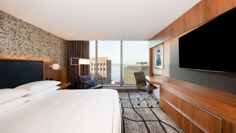 A modern hotel room with a large bed, a wall-mounted TV, a desk and chair, and a view of buildings and the water through wide windows.
