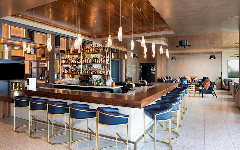 The image shows a modern bar with a central counter and bar stools, a well-stocked liquor display, stylish hanging lights, and seating areas.