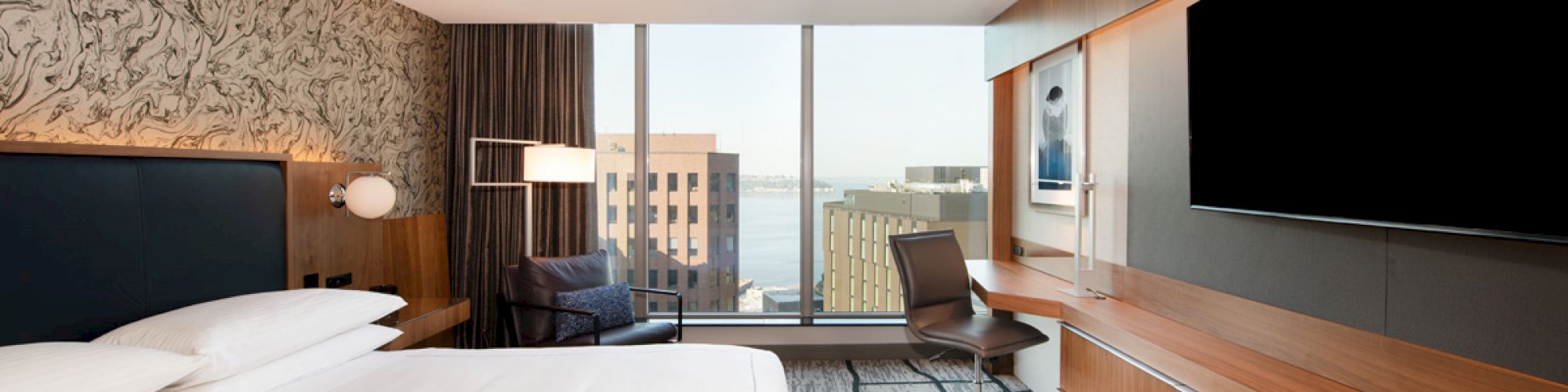 A modern hotel room features a large bed, wall-mounted TV, desk, chair, and a window with a city and water view outside.