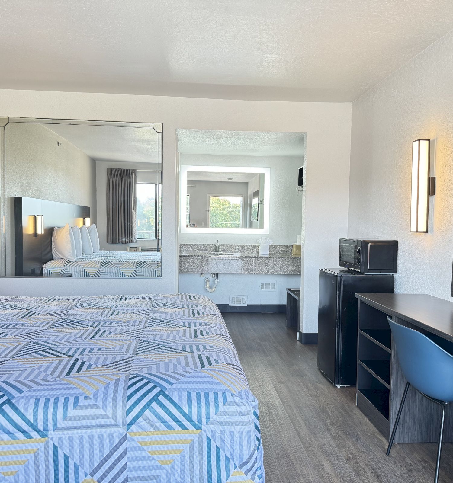 The image shows a modern hotel room with two beds, a large mirror, a desk with a chair, a microwave, a flat-screen TV, and light fixtures.