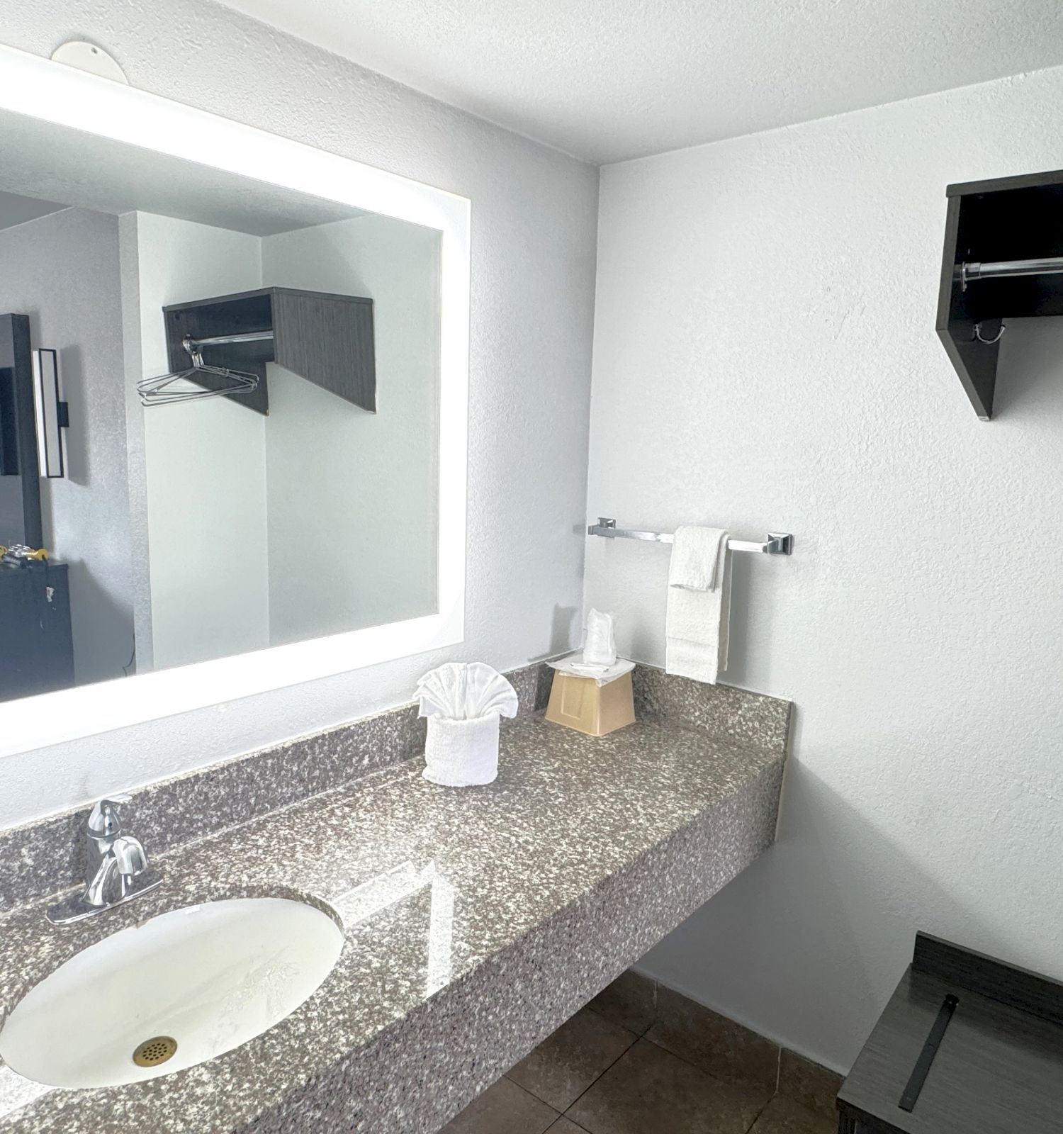 A modern bathroom with a granite sink, a large mirror with backlighting, a mounted towel rack, and a tissue box is shown in the image.