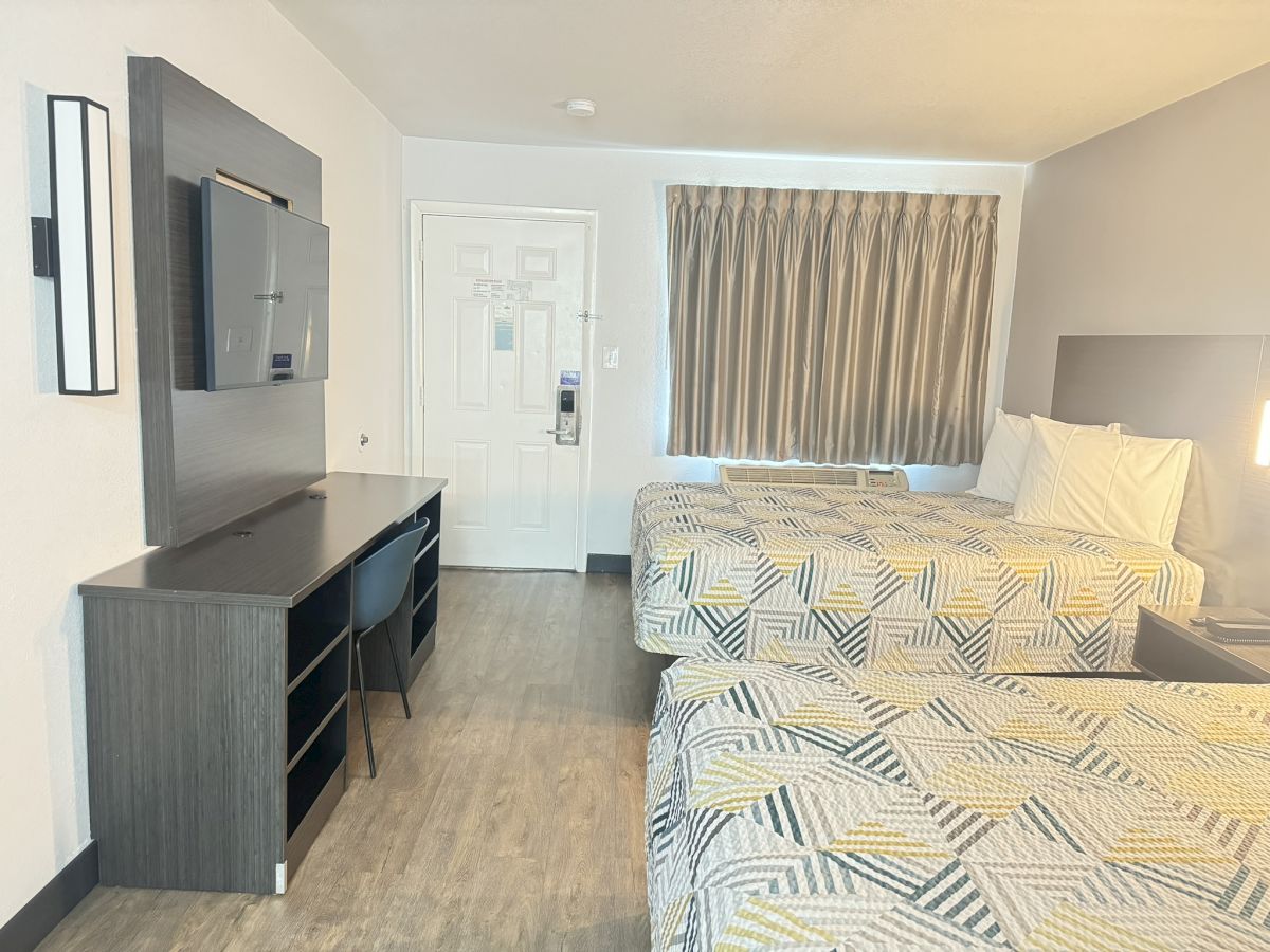 The image shows a hotel room with two beds, a desk with a chair, a wall-mounted TV, and a window with curtains. The decor is modern and minimalistic.