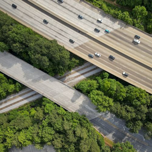 An aerial view of a highway with multiple lanes of traffic. Below the highway, a railway track and a road pass through a forested area.