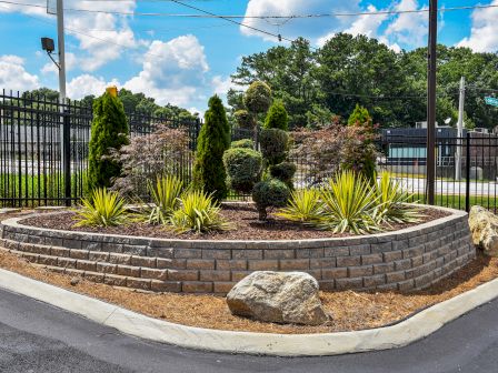 A landscaped garden bed features various plants, rock decorations, and a brick retaining wall in front of a black metal fence.