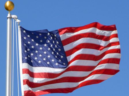 The image shows the flag of the United States of America waving on a flagpole against a clear blue sky.