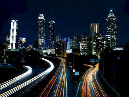 A cityscape at night showing illuminated skyscrapers and long-exposure light trails from vehicles on a multi-lane highway.