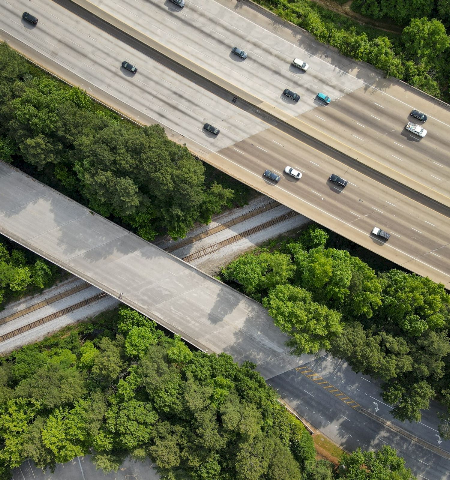 An aerial view of two highways with vehicles, surrounded by trees, and intersected by smaller roads and train tracks.